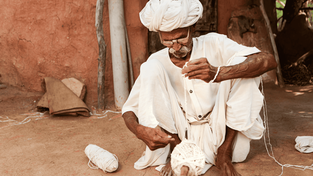 A-man-in-a-white-turban-sits-on-the-ground-in-Bishnoi-Village-India-surrounded-by-a-rural-landscape.png