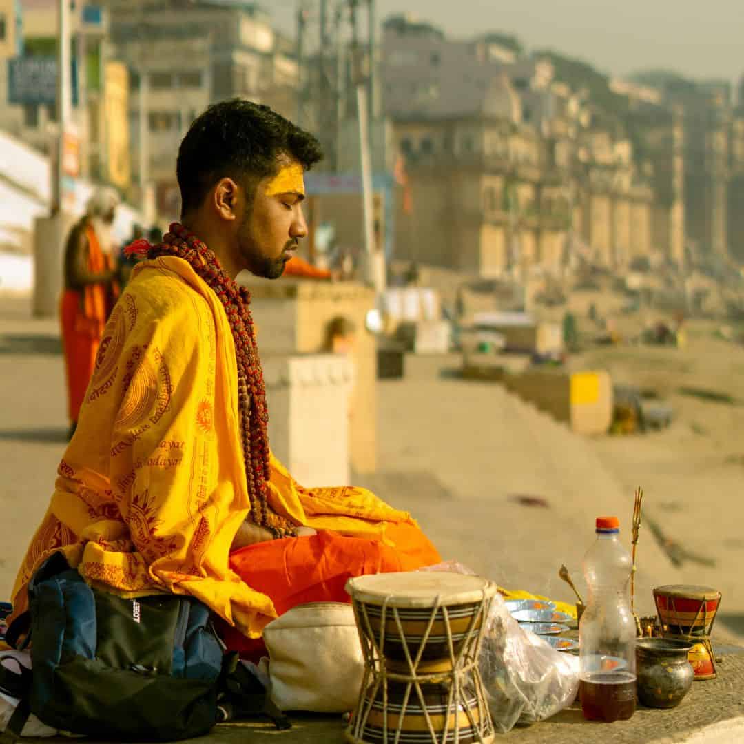 A man in a yellow robe sits by the riverbank in Varanasi, India, reflecting on the serene waters A man in a yellow robe sits by the riverbank in Varanasi India reflecting on the serene waters