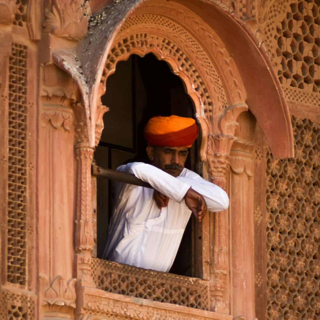 A man in an orange turban gazes out of a window, with the vibrant city of Jodhpur, India A man in an orange turban gazes out of a window with the vibrant city of Jodhpur India